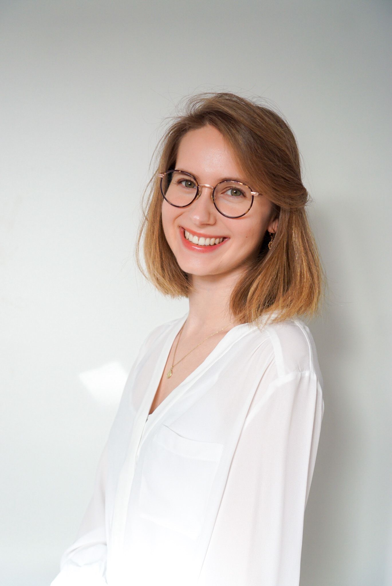 Portrait einer jungen Frau mit schulterlangem, hellbraunem Haar. Sie trägt eine runde Brille, eine weiße Bluse und eine feine goldene Halskette. Die Frau lächelt freundlich in die Kamera, der Hintergrund ist neutral und hell.